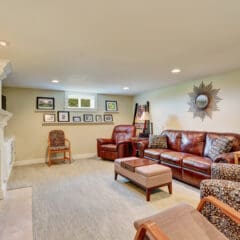 A cozy living room with beige walls, featuring a leather sofa, two armchairs with patterned upholstery, a wooden coffee table, and a fireplace. The wall above the fireplace has a display of framed photos, and a sunburst mirror hangs on the adjacent wall.