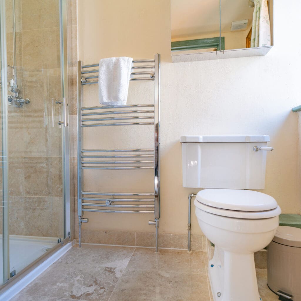 A clean, modern bathroom with a glass-enclosed shower, a white toilet, and a chrome heated towel rail with a white towel hanging on it. The floor and partially tiled walls are in a beige marble finish, and there's a mirror above the toilet.