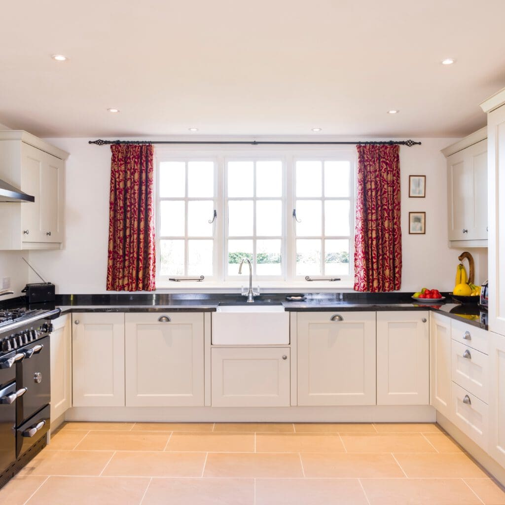 A bright kitchen with cream cabinets, black countertops, and a central white farmhouse sink beneath a large window with red patterned curtains. The kitchen features stainless steel appliances and light-colored tiled flooring. A bowl of fruit and a yellow kettle are on the countertops.