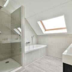 A contemporary bathroom with light grey tiles featuring a walk-in glass shower on the left, a bathtub with a handheld showerhead under a large skylight window in the center, and a white sink with a black cabinet on the right. The floor has light grey tiles.