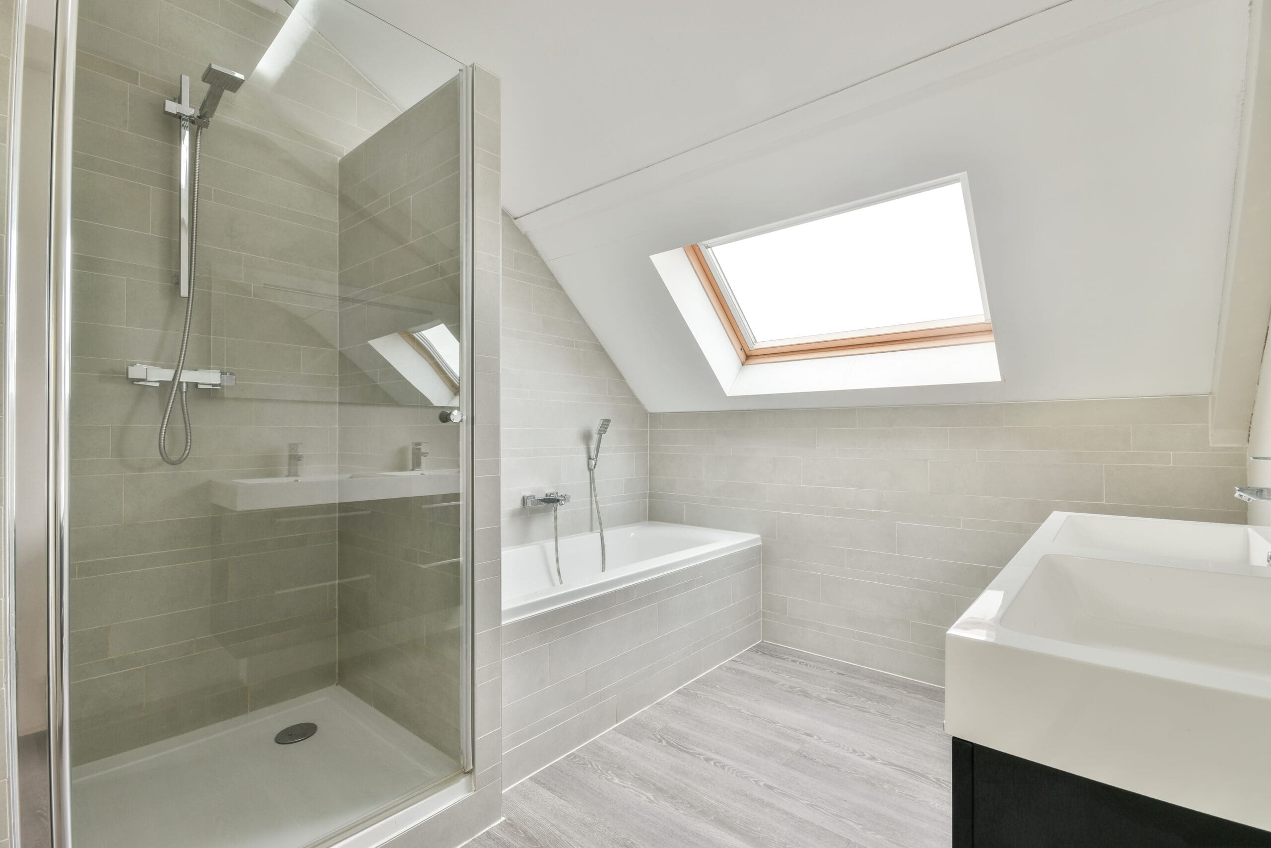 A contemporary bathroom with light grey tiles featuring a walk-in glass shower on the left, a bathtub with a handheld showerhead under a large skylight window in the center, and a white sink with a black cabinet on the right. The floor has light grey tiles.