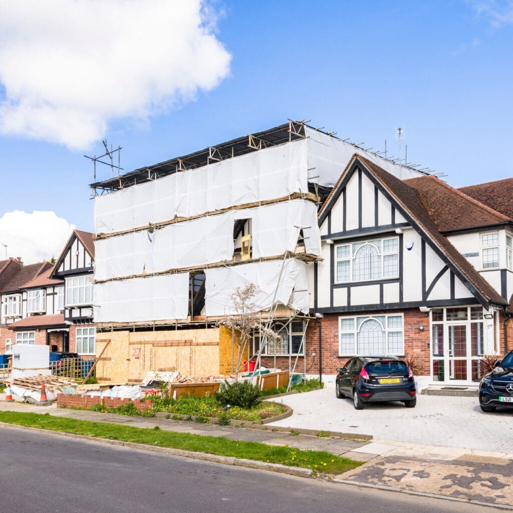 A large house with a Tudor-style facade is under renovation. The front of the house is covered with scaffolding and white sheeting. A blue and black car is parked in the driveway. The street in front has a grassy median. The sky is partly cloudy.