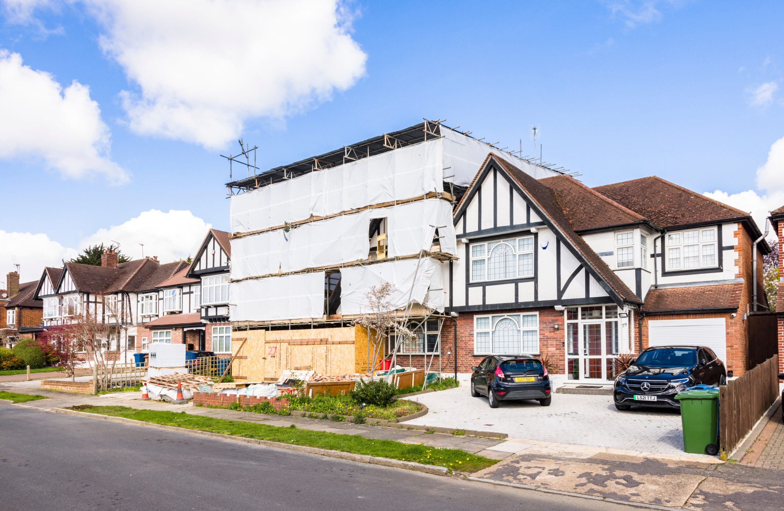 A residential street with a row of houses, one of which is under construction. The house under construction is covered in scaffolding and protective white sheeting. Several cars are parked in the driveway and on the street. The sky is clear with a few clouds.