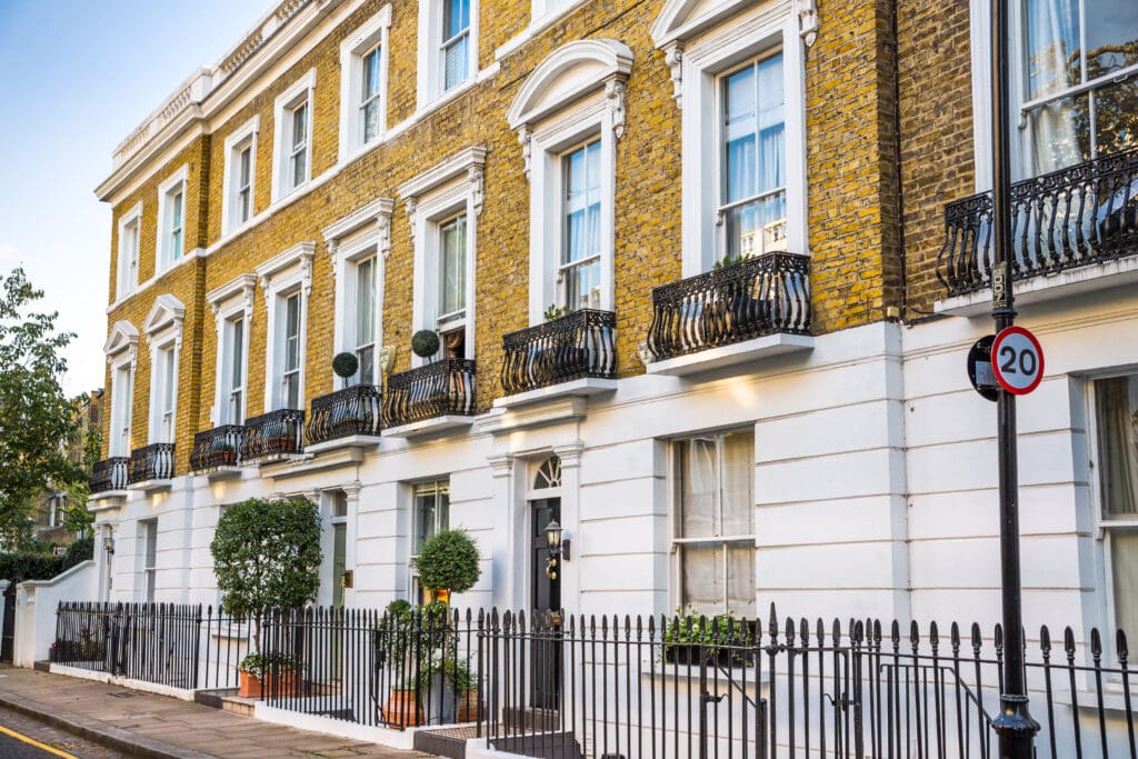 A row of classic, narrow, three-story townhouses with yellow brick facades, white trim, and black wrought iron balconies. There are neatly trimmed topiary plants in front of the buildings and a 20 mph speed limit sign on the right. .