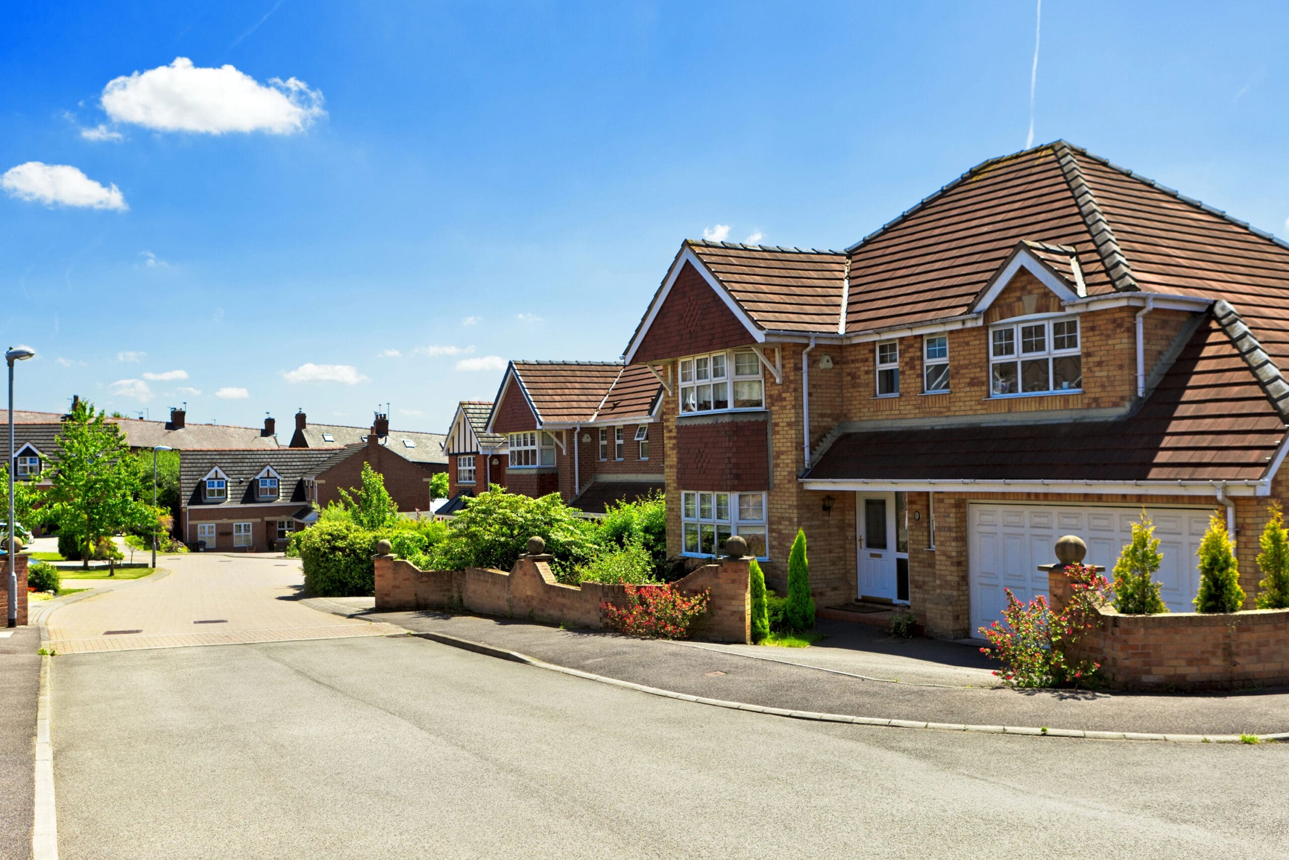 A suburban street lined with modern brick houses featuring tiled roofs and large windows on a sunny day. The houses have well-manicured gardens and driveways, with a clear blue sky in the background.