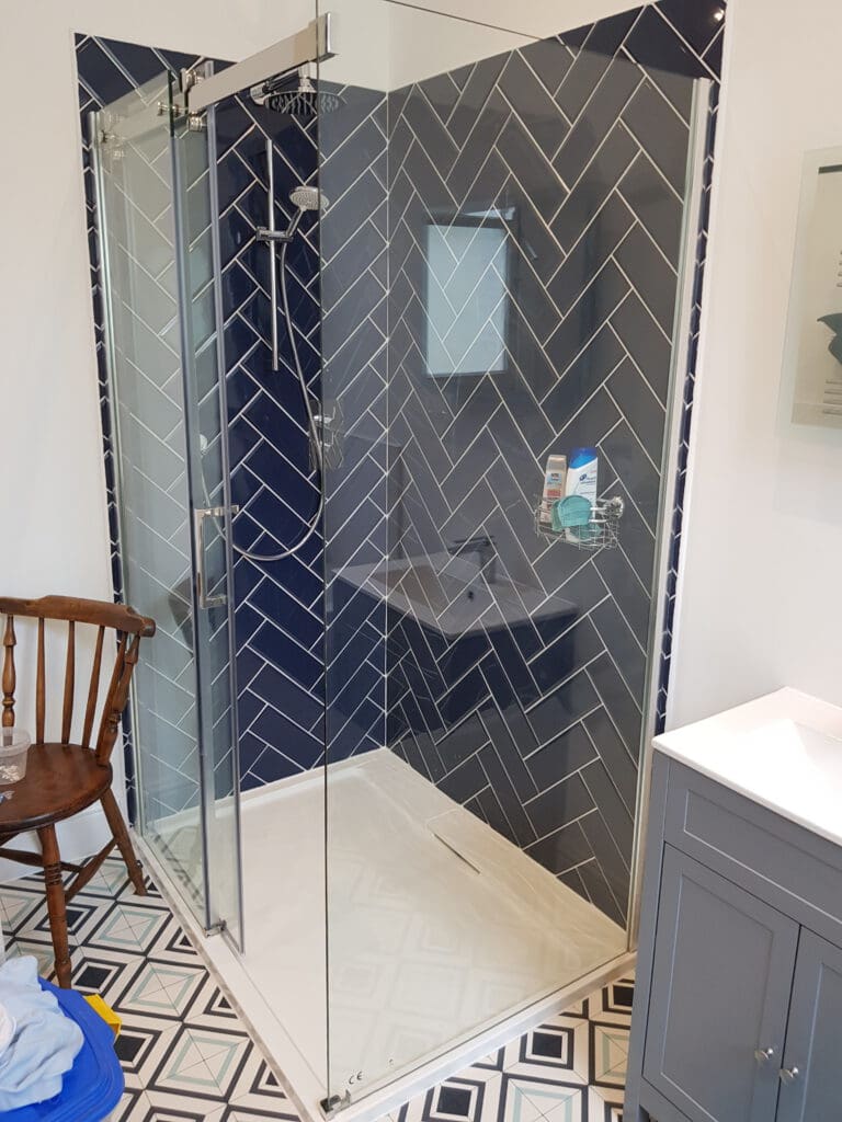 Modern bathroom shower with dark herringbone tile design, glass door, and chrome fixtures. A wooden chair is placed outside the shower, and part of a gray vanity is visible on the right. Unique geometric patterned floor tiles add a distinct touch to the decor.