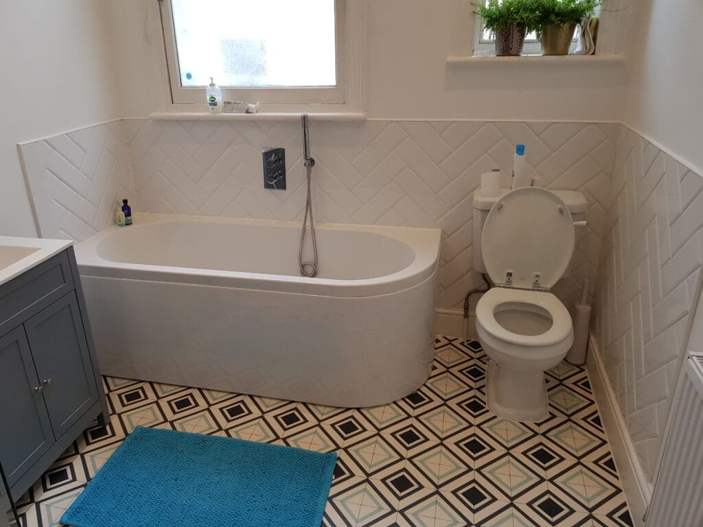 A bathroom with a modern white bathtub, toilet, and gray vanity. The walls have white tiles arranged in a herringbone pattern. Decorative plants sit on a ledge above the toilet. The floor has black and white geometric tiles, and there is a blue bath mat on the floor.