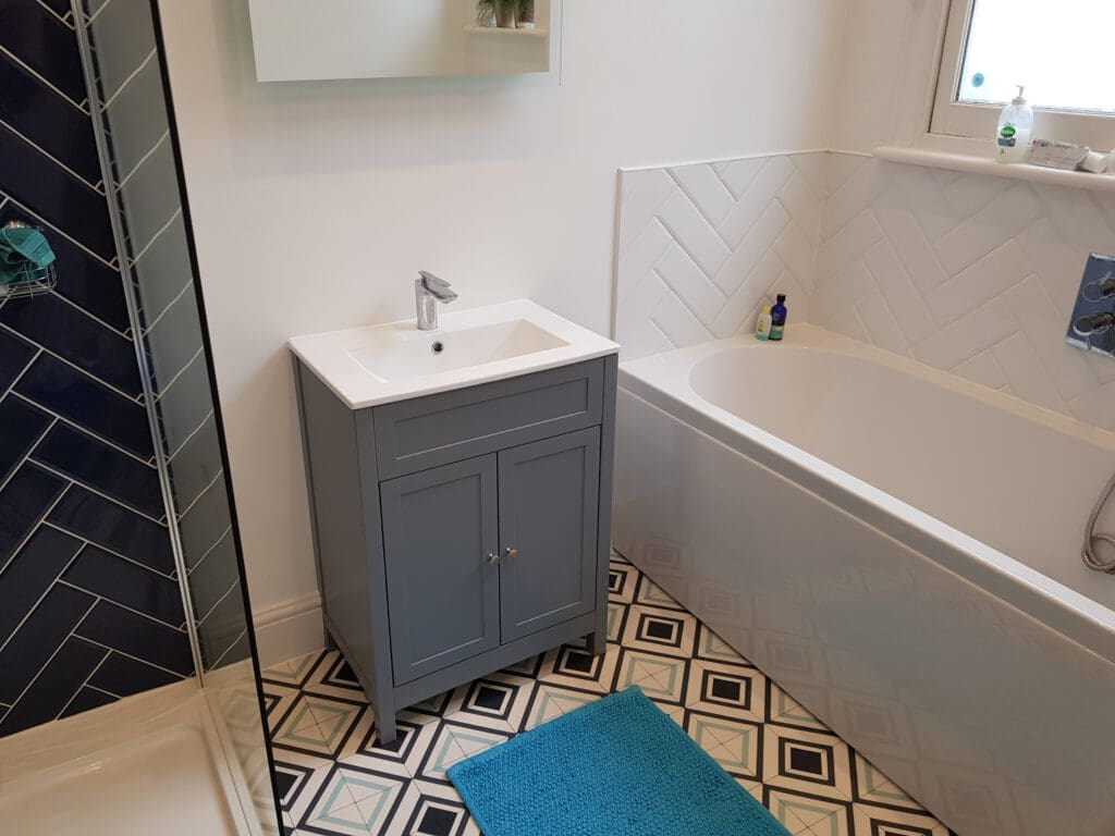 A modern bathroom featuring a grey vanity with a white sink, a bathtub adjacent to it, and a blue mat on geometric patterned floor tiles. The shower area has dark blue wall tiles with a glass door. The walls are white, and a window is near the bathtub.