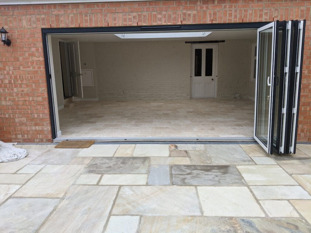 A spacious, empty garage with beige tiled flooring and a large, open, folding door that leads to a patio with stone tiles. The garage walls are painted white, and there is a white door in the back wall. The exterior wall is made of red bricks.