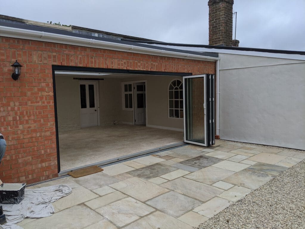 A partially open bi-fold door leads to an indoor area in this outdoor patio scene. The patio features light-colored stone tiles and is bordered by brick and plastered walls. Two white interior doors and a large arched window are visible inside the room.