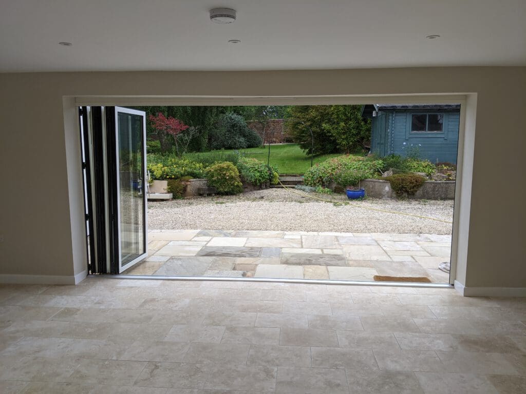 A spacious room with large bi-fold glass doors opening to a patio with stone tiles. Beyond the patio, there's a garden area featuring bushes, flowers, and a small green shed in the right corner. The interior floor is cream tiled.