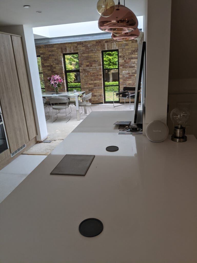 A modern kitchen with a long white countertop, wooden cabinets on the left, and rose gold hanging lights. In the background, there's a dining area with a table, chairs, and a vase of pink flowers, next to floor-to-ceiling windows showing greenery outside.