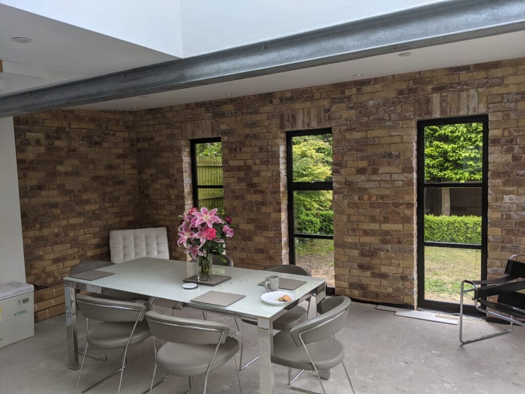 A modern dining room with exposed brick walls and tall windows. A glass table is surrounded by grey chairs and adorned with a vase of pink flowers. Outside the windows, greenery is visible. The room features a white ceiling and concrete floor.