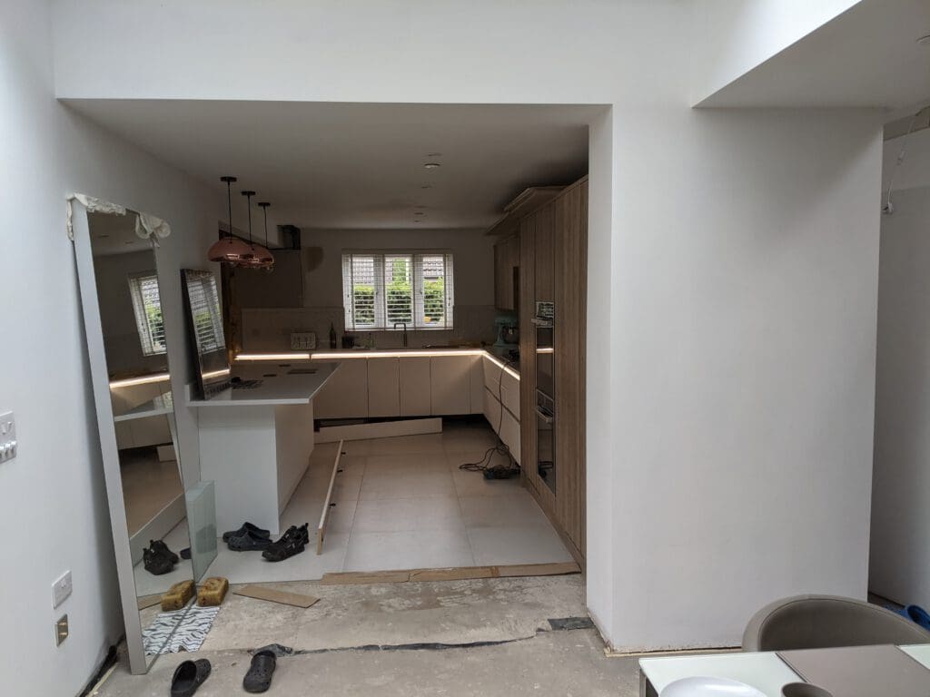 Image of a modern, partially furnished kitchen with white cabinets, a light wooden shelving unit, copper pendant lights, and a large window with white shutters. A pair of shoes and a ladder are visible on the unfinished floor, indicating ongoing construction.