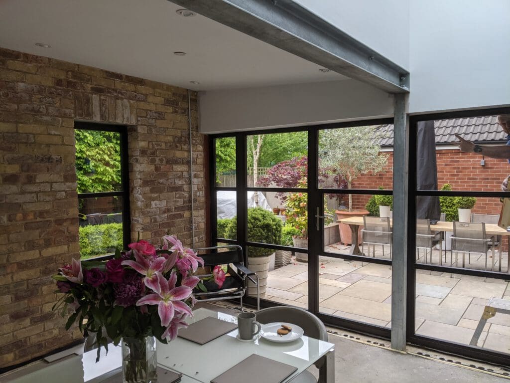 A modern dining area with large windows. The room features exposed brick walls, a table set for breakfast with a coffee cup and a plate, and a vase with pink lilies. Outside, there is a patio with a dining set, plants, and a view of a brick building.