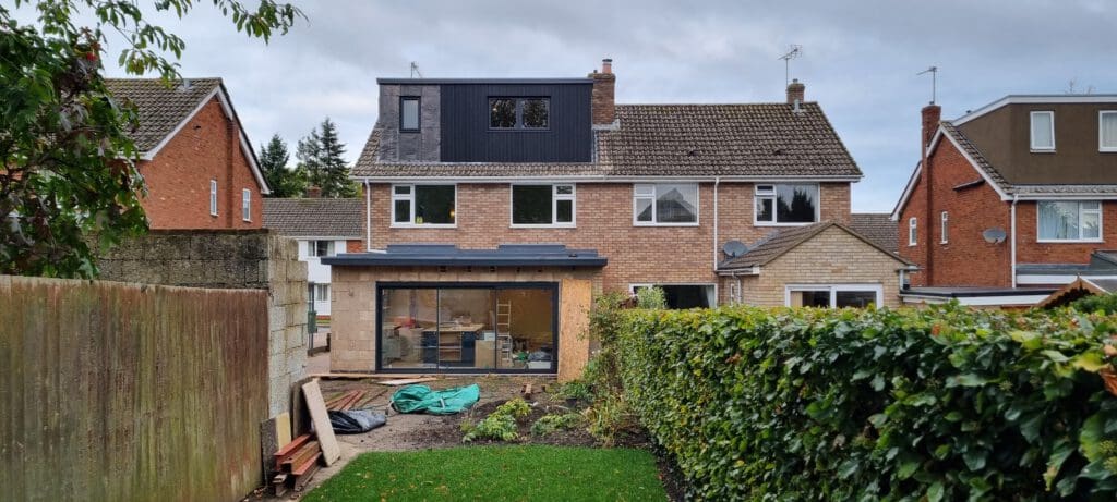 A two-story house with a gray dormer extension on the roof and an additional rear extension with large windows. The backyard is partially landscaped with a green lawn, hedges, and some construction materials scattered around. Neighboring houses are visible on both sides.