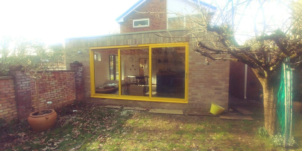 A small, modern house extension with brick walls and large yellow-framed sliding glass doors. Inside, a table and chairs are visible. Outside, there is a garden with fallen leaves, a potted plant, a leafless tree, and a yellow bucket on the ground.