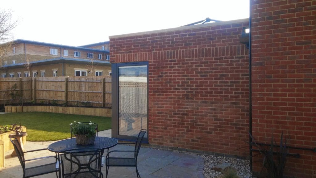 A brick building with a large glass door is adjacent to a paved patio area featuring a round metal table and two chairs. The patio overlooks a fenced lawn with some plants around the edges. Another building is visible in the background.