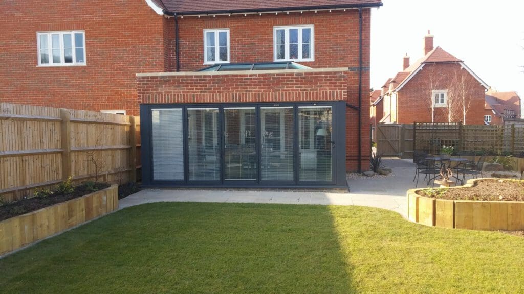 A spacious backyard with well-maintained grass, bordered by wooden planters. A modern extension with large glass doors is attached to a brick house. A small patio area with a table and chairs is seen on the right, leading to a wooden fence and neighboring houses.