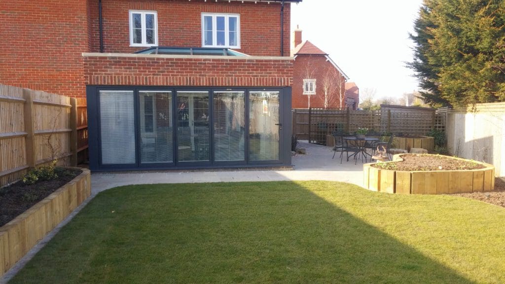 A backyard with a green lawn, wooden raised garden beds, a patio with a round table and chairs, and a modern red-brick house with large sliding glass doors in the background. The yard is enclosed by wooden fences and tall hedges.