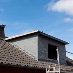 A partially constructed dormer on a gray tiled roof under a clear blue sky. There is scaffolding on the right and a ladder leaning against the roof. A chimney is visible to the left of the dormer.