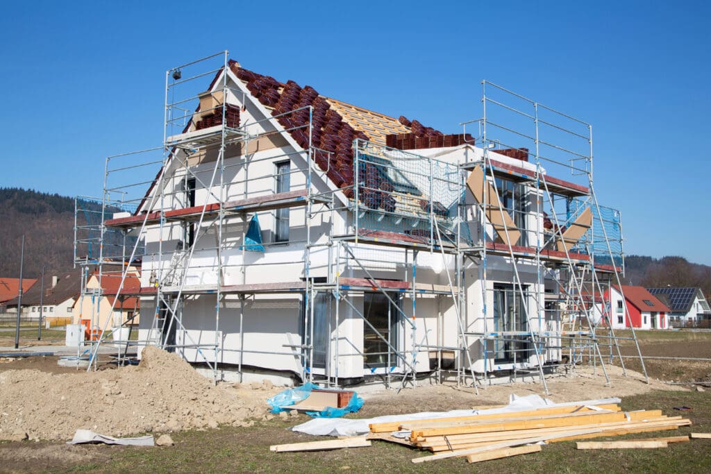 A two-story house under construction with scaffolding surrounding it. The roof is partially covered with tiles, and construction materials are scattered around the yard. The sky is clear and blue, indicating a sunny day. Several other houses are visible in the background.