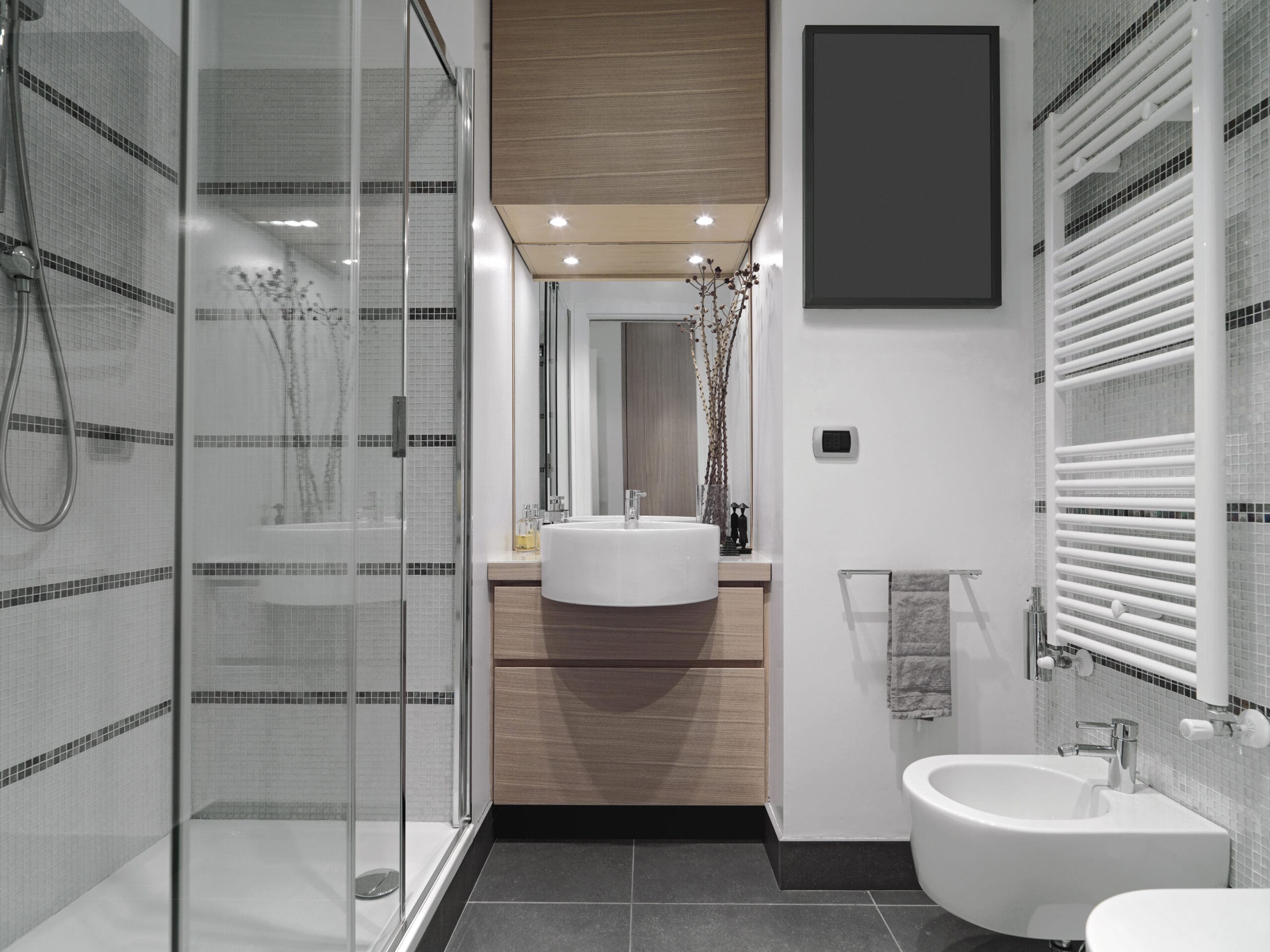 Modern bathroom with a glass-enclosed shower on the left, a floating wooden vanity with an oval sink and large mirror at the center, and a bidet next to a towel warmer on the right. The walls have a gray and white tile design, and the floor is dark-colored.