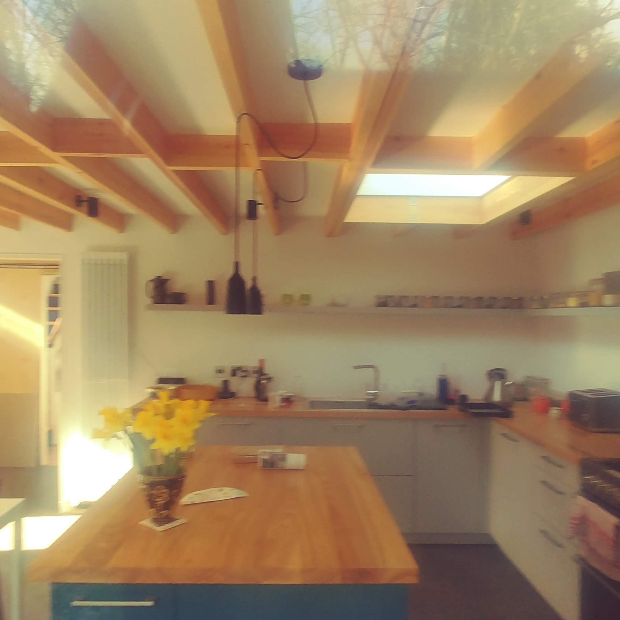 A modern kitchen with wooden beams and white walls. A large wooden island sits in the center, adorned with a vase of yellow flowers. Shelves with jars line the back wall, and a skylight lets in natural light. The space is tidy with minimalistic decor.