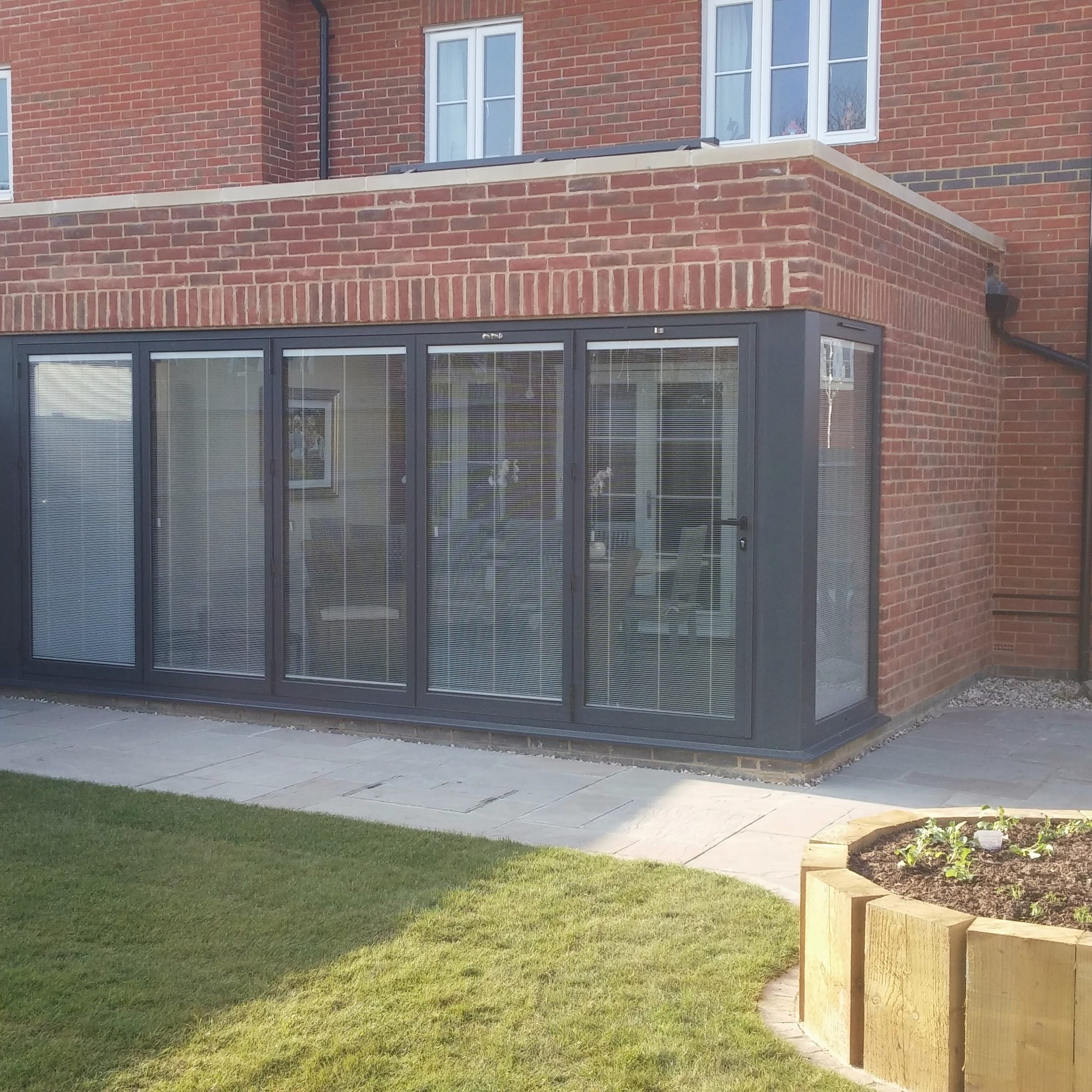 A modern brick house with a large glass-enclosed extension featuring floor-to-ceiling windows and blinds. The extension overlooks a neatly landscaped backyard with a grassy lawn, a raised flower bed, and a small patio area with chairs and a table.