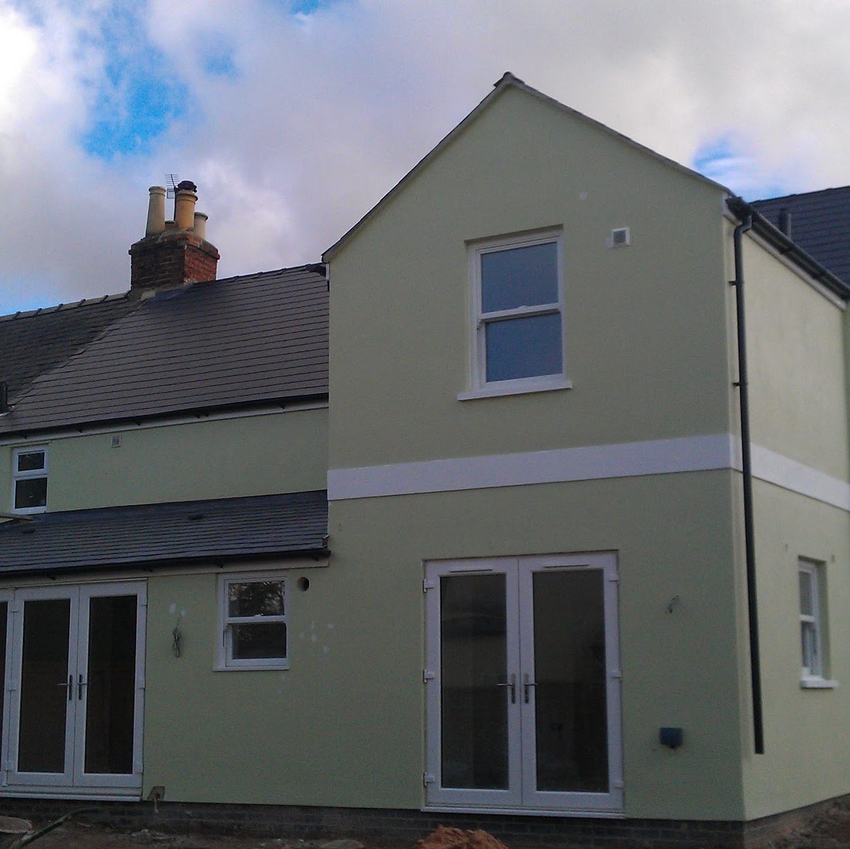 A light green two-story house with a gray roof and white trim around the windows and doors. The house has multiple large windows, including two sets of French doors on the ground floor. The sky is partly cloudy. The house has a small yard under construction.