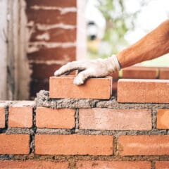 A close-up view of a construction worker's arm and hand wearing a glove, placing a brick on a wall being built. The partially built wall is made of red bricks with visible mortar between them. The background is blurred, focusing on the worker's task.