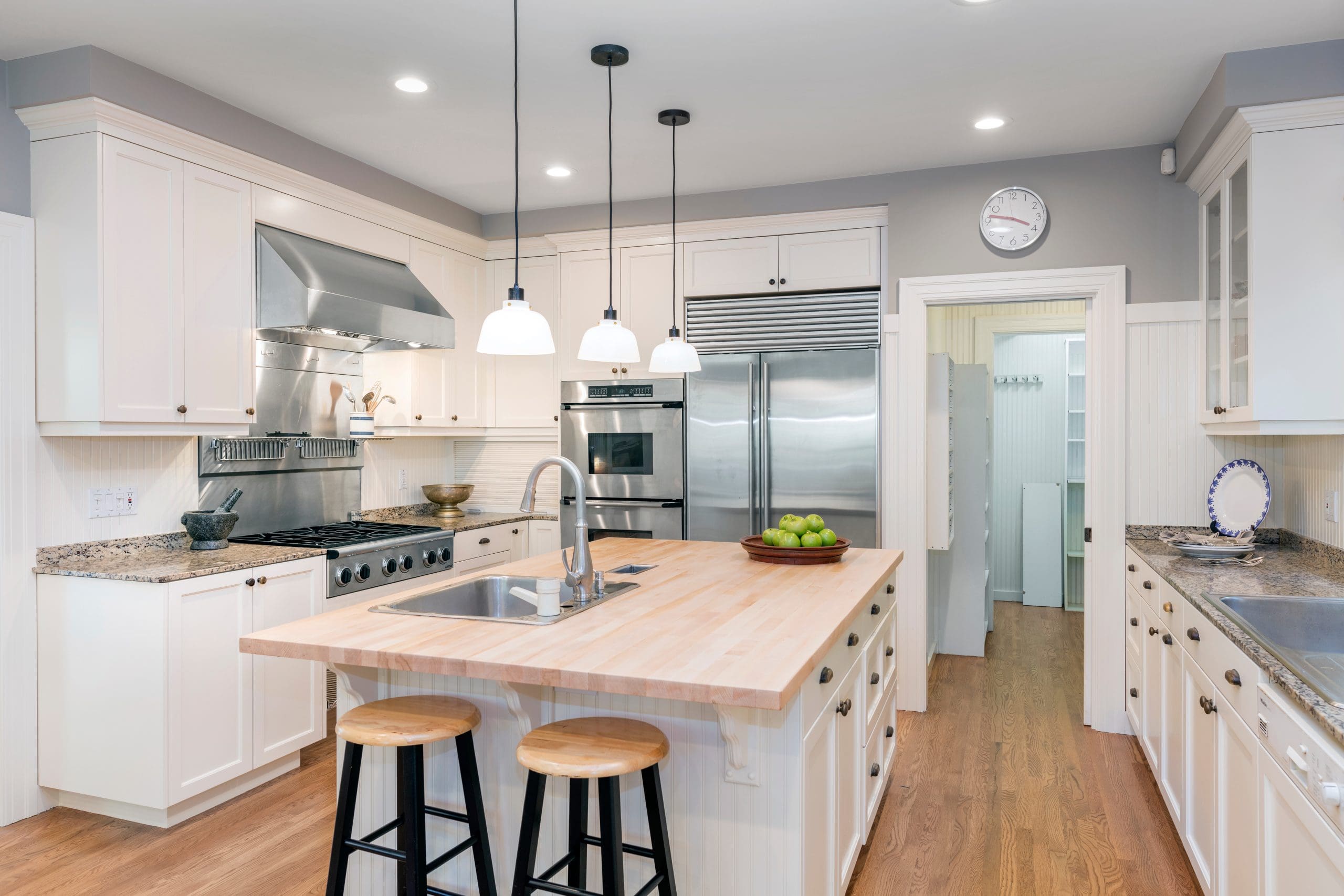 A modern kitchen with white cabinets, a wooden island with two stools, stainless steel appliances including a double fridge and a stove, and granite countertops. Hanging pendant lights illuminate the island, and a bowl of green apples sits atop it.