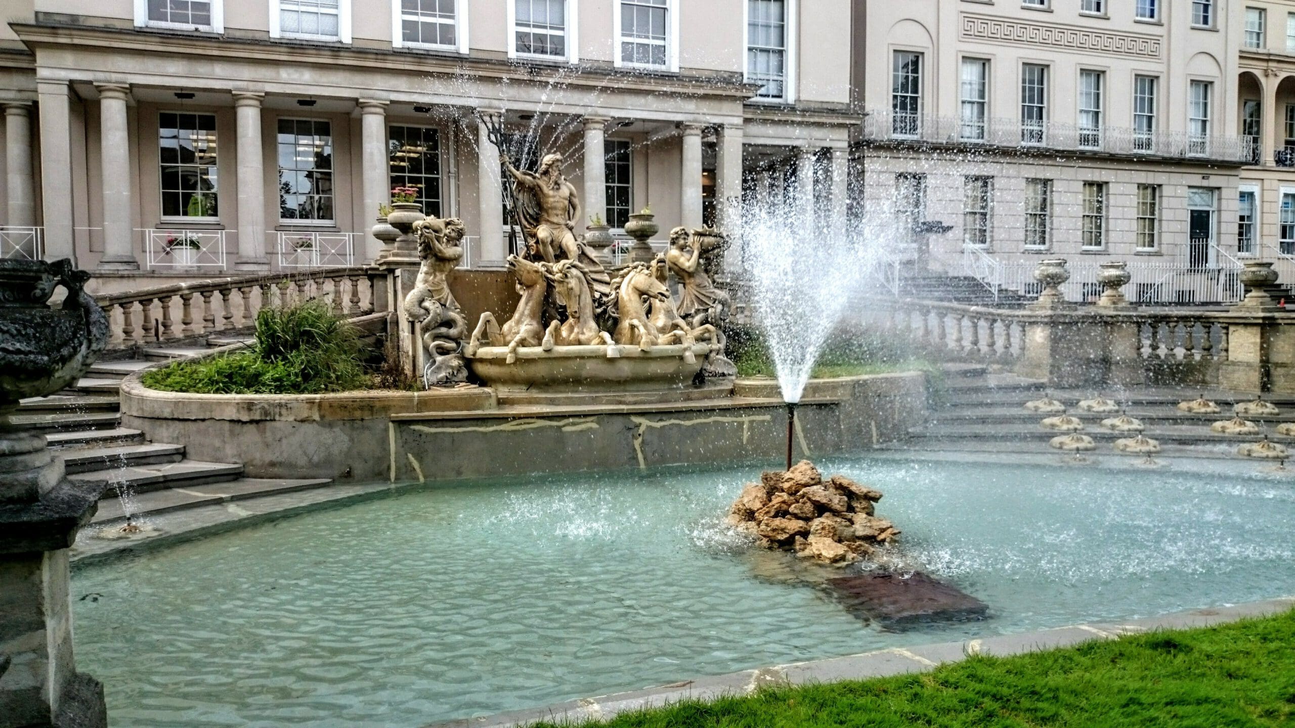 A decorative fountain with statues, featuring Neptune and sea creatures, stands in front of a historic building with columns. Water cascades from the statues and a central jet, creating a serene pool surrounded by manicured greenery.