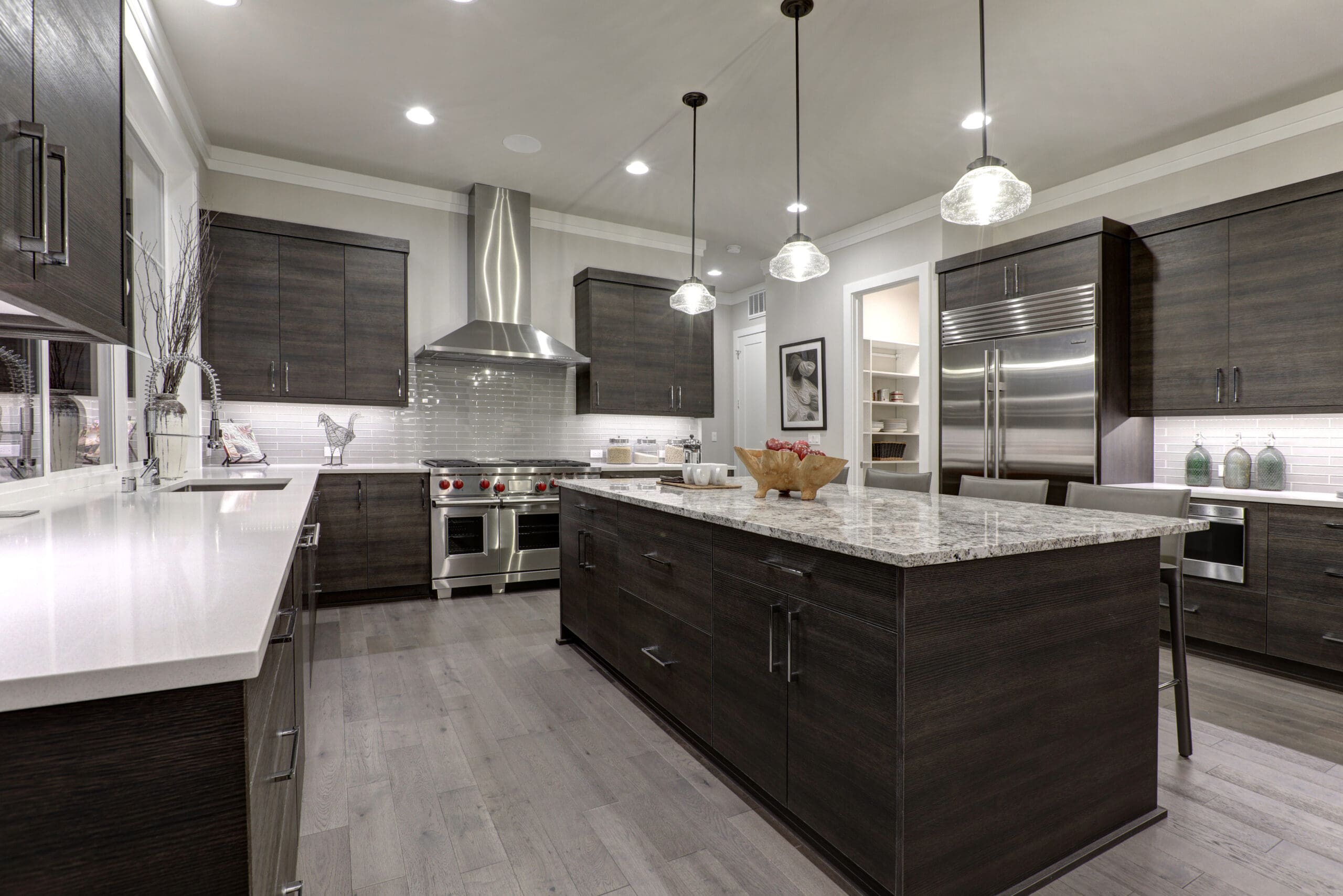 Modern kitchen with dark wood cabinets, stainless steel appliances, and a large island with granite countertop. The room is well-lit with recessed lighting and three pendant lights above the island. The backsplash is white subway tile, and the floors are light wood.