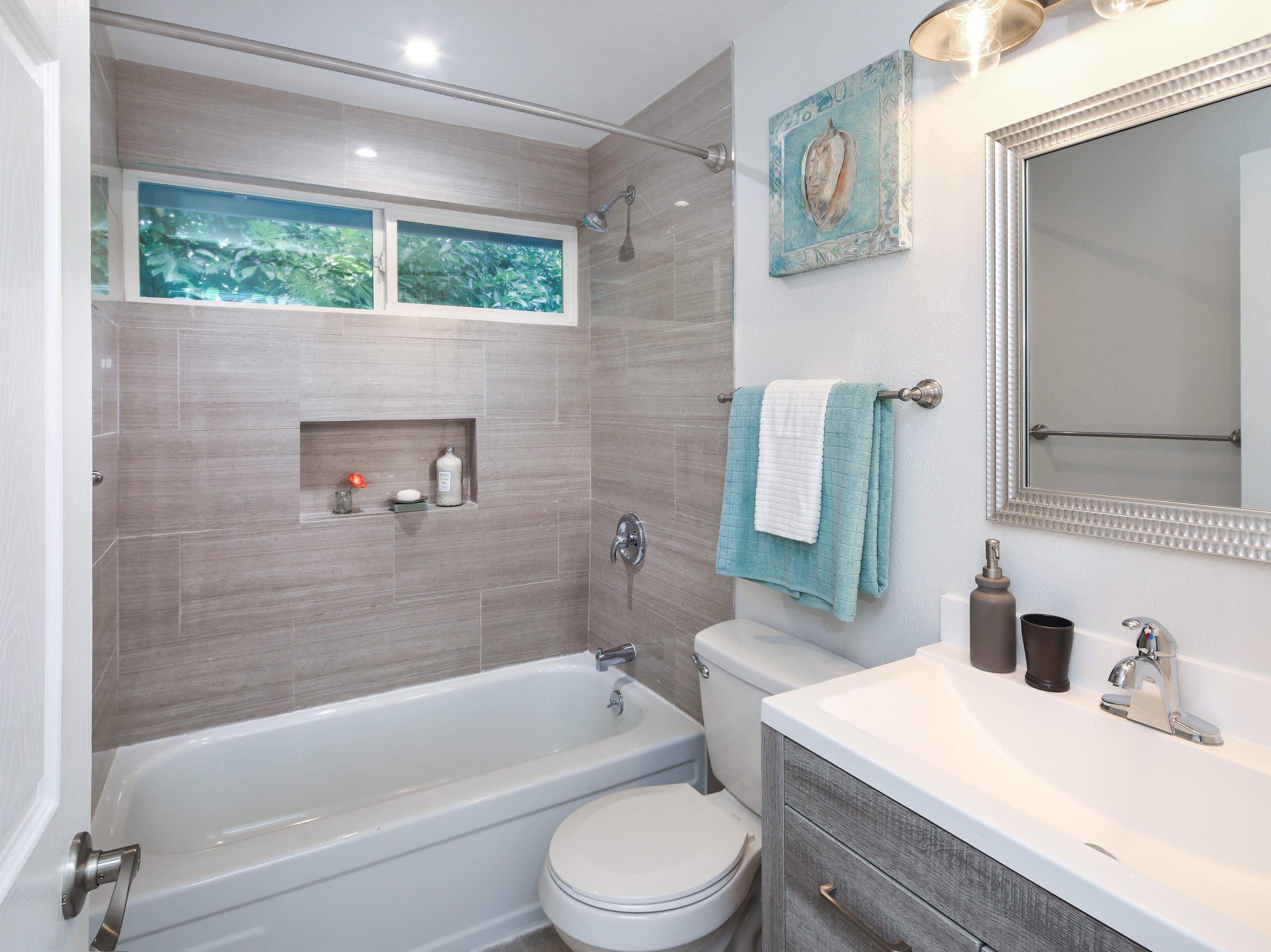 A modern, well-lit bathroom with gray tiled walls, a white bathtub, and a white toilet. Above the toilet is a towel rack with teal towels. A rectangular mirror and light fixtures are above the white sink on the right. A framed artwork hangs on the wall.