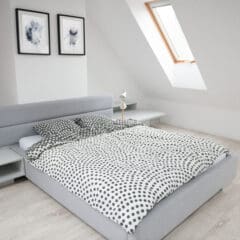 A minimalist bedroom with a grey upholstered bed against a white wall. The bed is made with white and black polka dot bedding. There are two black and white framed artworks above the bed, and a skylight window on the slanted ceiling to the right, illuminating the room.