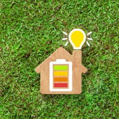 A small wooden house silhouette with a colored battery icon (green, yellow, orange, red) on its side and a glowing light bulb on its chimney, placed on a grassy surface.