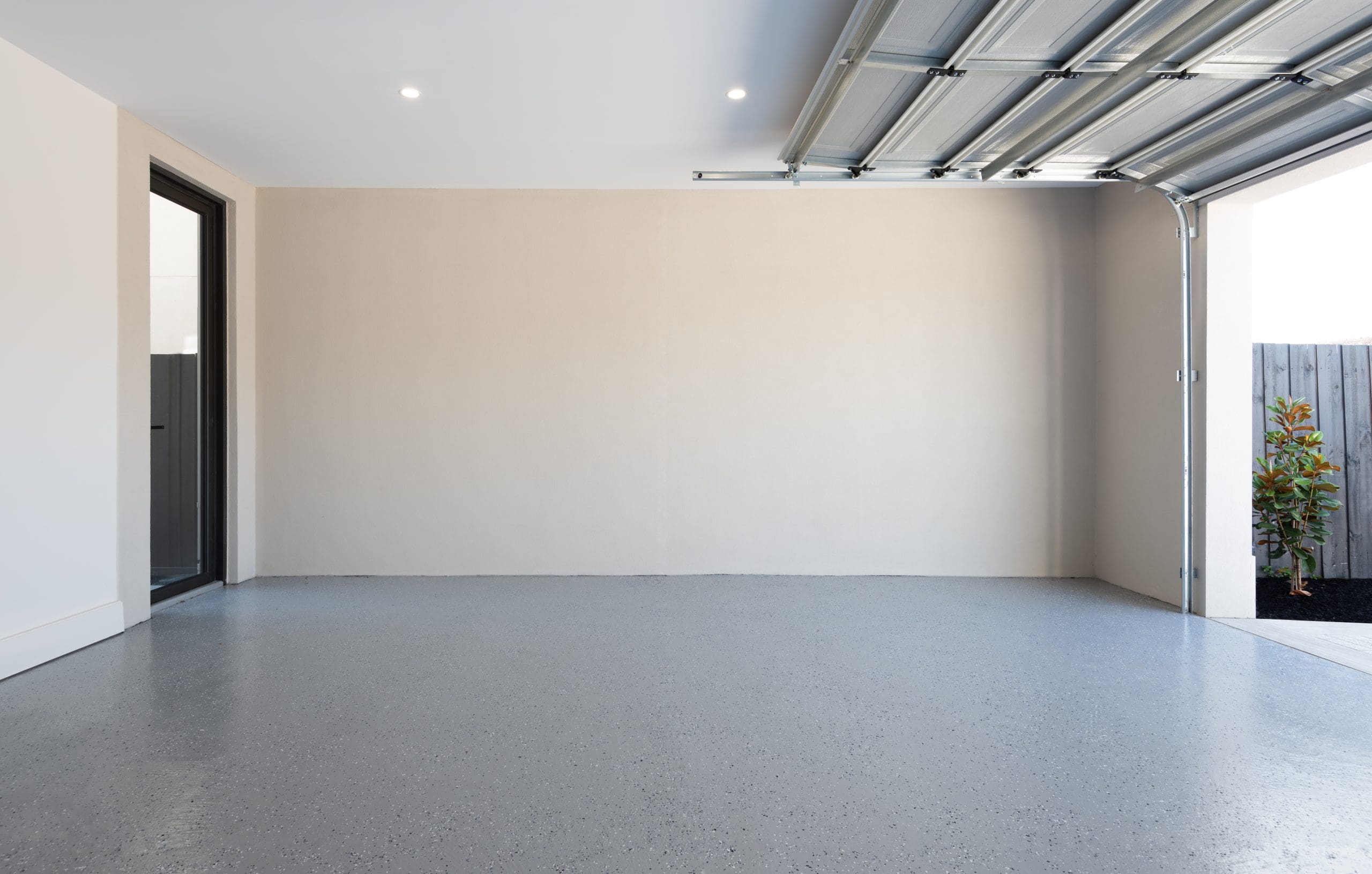 An empty, clean garage with light grey walls and a polished grey concrete floor. The garage door is partially open, revealing some plants and a wooden fence outside. A glass door on the left leads into the house. The space is well-lit with recessed ceiling lights.