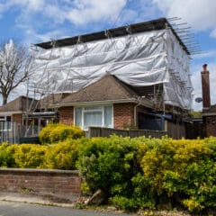 A house undergoing construction is covered with scaffolding and plastic sheeting. The surrounding yard has green foliage and yellow flowers. Another house is visible to the left, and a leafless tree is in the background against a partly cloudy sky.