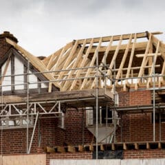 A partially constructed brick house with visible wooden roof trusses and scaffolding surrounding the structure. The roof framework is unfinished, and boards and materials are scattered at the construction site. The sky is overcast.