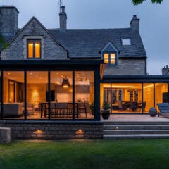 A modern house at dusk features large glass windows illuminating the interior with warm light. The exterior comprises stone walls with a dark shingled roof. The backyard includes a lush green lawn, a patio area with seating, and soft landscape lighting.