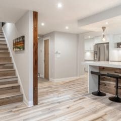 A modern basement with light wood flooring, a staircase to the left, a kitchen area to the right featuring a white countertop with two black bar stools, pendant lights, stainless steel appliances, and a potted plant. The space is well-lit with recessed lighting.