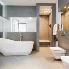 A modern bathroom with sleek gray tiles on the walls and floor. It features a white standalone bathtub, a wall-mounted toilet and bidet, large mirrors, recessed lighting, and a walk-in shower with a glass accent wall. Towels are hung beside the shower.