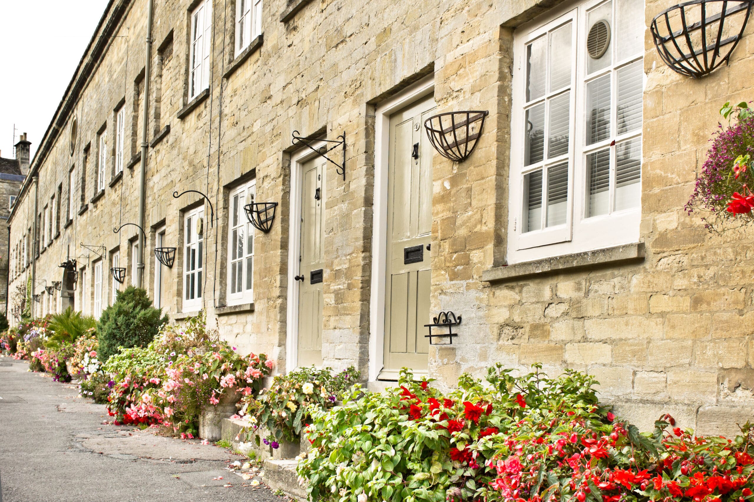 A row of charming stone houses with white windows and doors is adorned with colorful flower baskets and pots. A paved sidewalk runs along the front of the houses, and the overall scene is bright and inviting, showcasing a vibrant display of greenery and flowers.