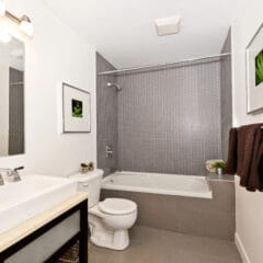 A modern bathroom with a white sink and vanity on the left, a large mirror above, a toilet in the center, and a bathtub with a showerhead on the right. The walls feature gray tiles and white paint, with framed plant art and brown towels for decor.