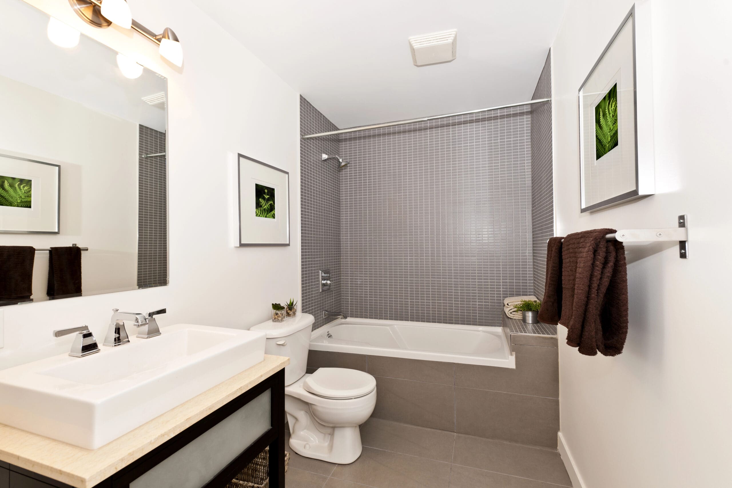 A modern bathroom with a white sink and vanity on the left, a large mirror above, a toilet in the center, and a bathtub with a showerhead on the right. The walls feature gray tiles and white paint, with framed plant art and brown towels for decor.