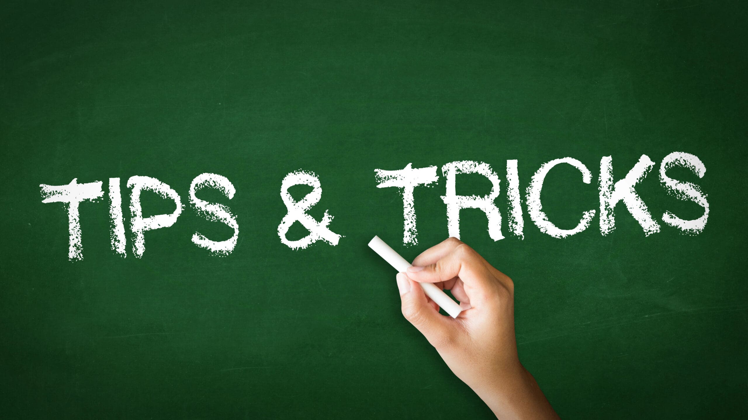 A hand holding a piece of chalk writes "TIPS & TRICKS" on a green chalkboard in white capital letters. The background is entirely green, resembling a school chalkboard.