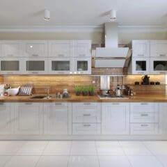 A modern kitchen with white cabinetry, a wooden countertop, and a glossy tile backsplash. The kitchen features a stainless steel range hood, an induction cooktop, and various kitchen utensils and appliances, including a kettle and a coffee maker, neatly arranged.