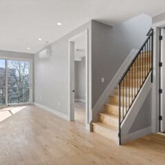 A modern, empty living space with light wood floors, gray walls, and recessed lighting. The room features a staircase with black railings, a sliding glass door leading to a balcony, and ample natural light streaming in from the large window.