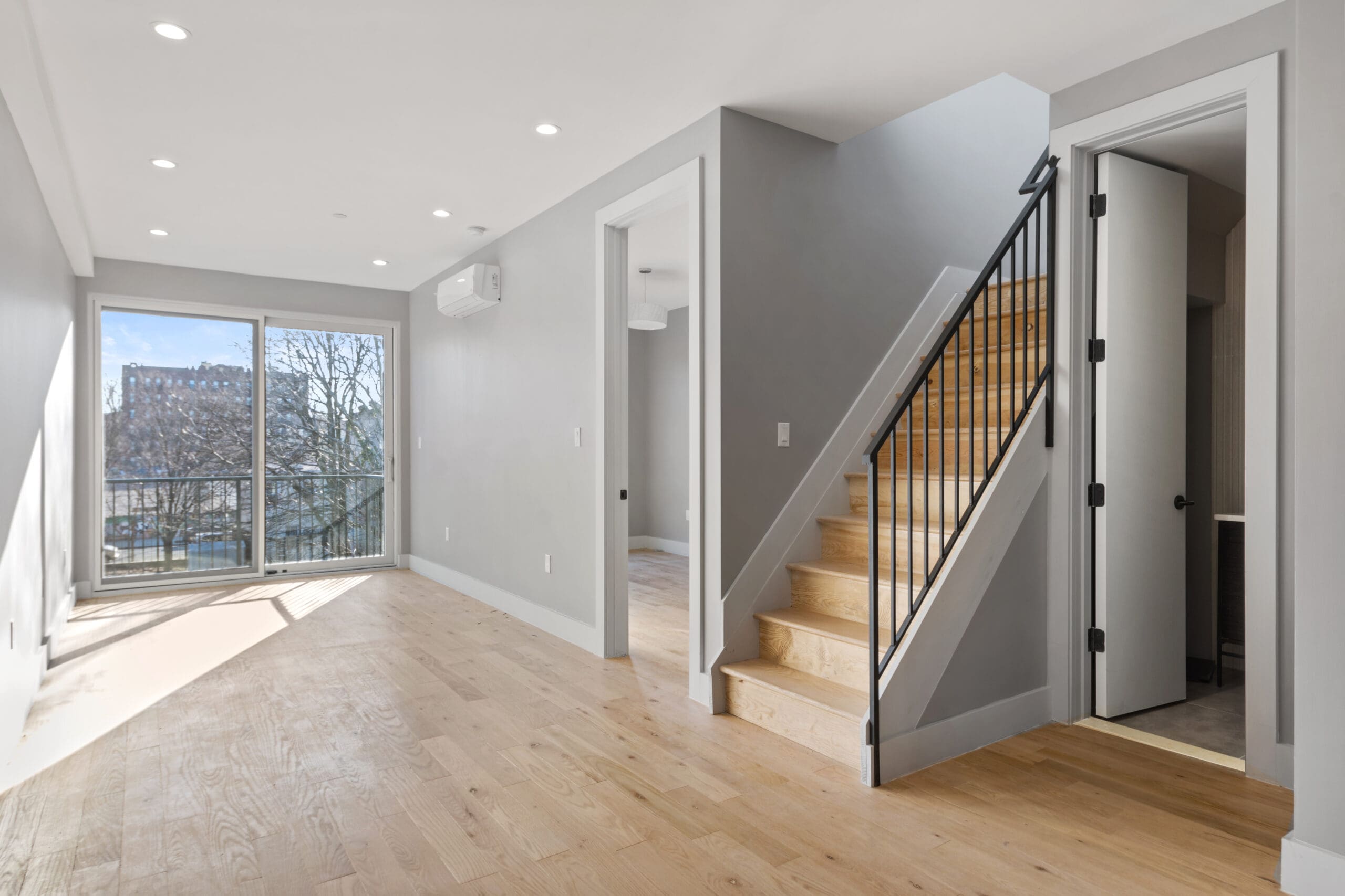 A modern, empty living space with light wood floors, gray walls, and recessed lighting. The room features a staircase with black railings, a sliding glass door leading to a balcony, and ample natural light streaming in from the large window.